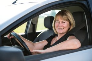 Nainen istuu auton ratissa ja hymyilee